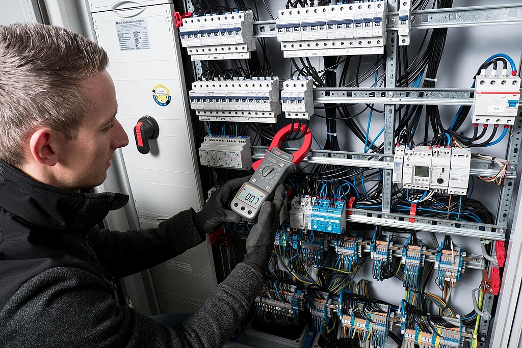 Elektrotechniker prüft in einem Schaltschrank Leitungen mit einem Zangenstrommesser