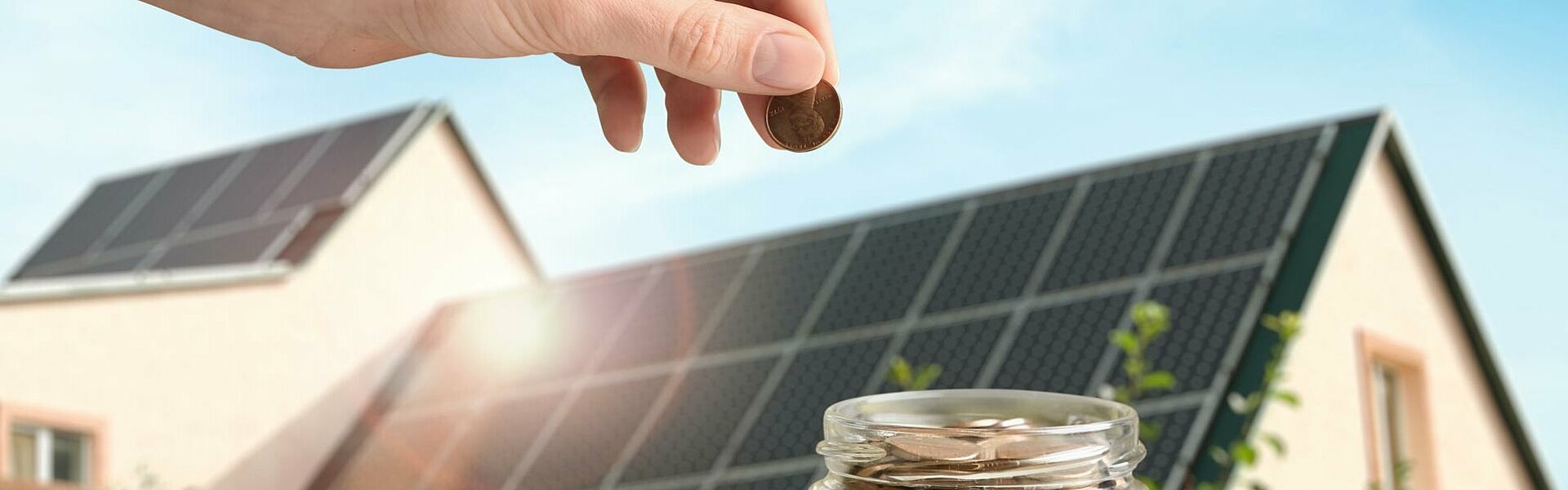 Mit Münzen gefülltes Glas vor einem mit Photovoltaikmodulen bestückten Dach eines Privathaushalts